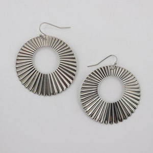 May include: A pair of silver hoop earrings with a textured, sunburst design. The earrings are large and circular.