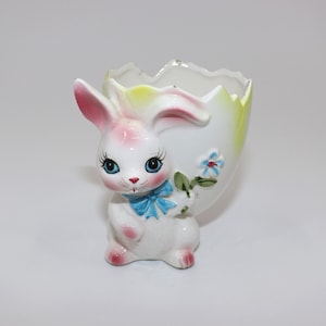 May include: A white ceramic Easter egg holder shaped like a bunny. The bunny has pink ears, blue eyes, and a blue bow. The egg holder has a yellow and green flower on the side.