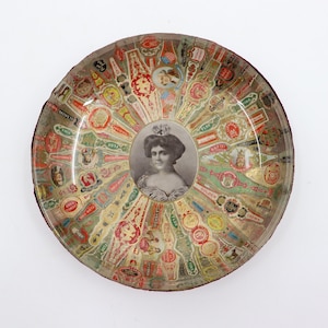 May include: A round, glass ashtray featuring a colourful collage of cigar labels. The labels display various brand names, such as "Golden Crown", "La Verde", and "Flor Fira". A portrait of a woman in a white dress is at the centre of the ashtray.