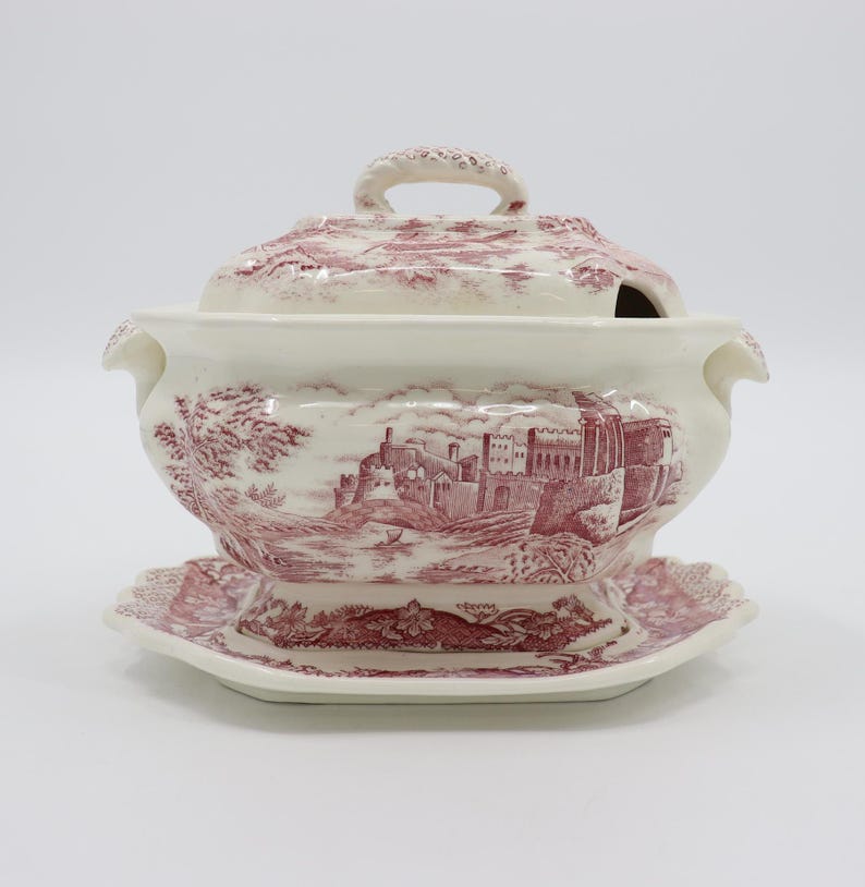 May include: A white ceramic gravy boat with a red and white castle design. The boat has a lid and a matching base.