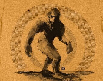 May include: A mustard yellow t-shirt with a black and white graphic of Bigfoot standing in a circle of concentric rings.