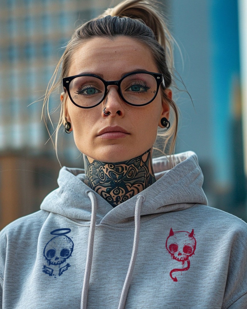May include: A gray hoodie with a black and white skull with angel wings on the left side and a red skull with devil horns and a tail on the right side. The model is wearing the hoodie and has a black nose ring and black glasses.