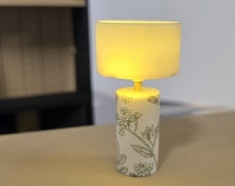 May include: A white lampshade with a floral patterned base. The lamp is turned on and the light is illuminating the room.