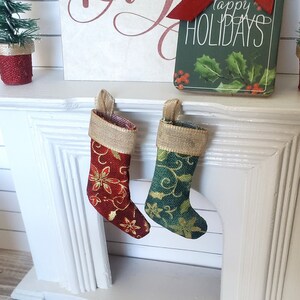 May include: Two miniature Christmas stockings hanging on a white mantel. The stocking on the left is red with gold accents and the stocking on the right is green with gold accents. The stockings are decorated with a floral pattern.