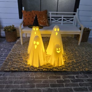 May include: A Halloween-themed display featuring four glowing ghost figures of varying sizes. One ghost wears a black witch hat. The ghosts are illuminated from within, casting a warm, inviting glow. The scene is set on a rug with a bench and pillows in the background.