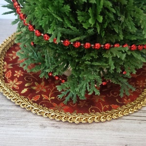 May include: A small artificial Christmas tree with red bead garland, sitting on a red and gold patterned tree skirt with a gold trim.