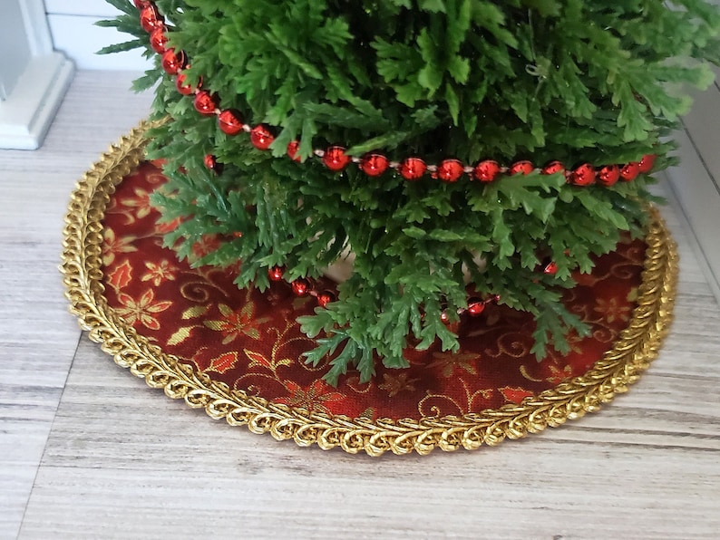 May include: A small artificial Christmas tree with red bead garland, sitting on a red and gold patterned tree skirt with a gold trim.