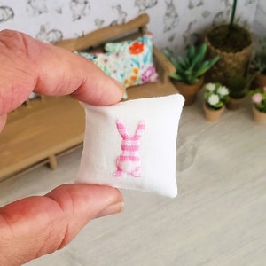May include: A white pillow with a pink and white striped bunny embroidered on it. The bunny is facing away from the viewer and has a fluffy tail.