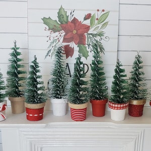 May include: Seven miniature artificial Christmas trees in pots with red and white ribbon accents. The trees are green and have a snowy white finish. The trees are arranged on a white mantel.