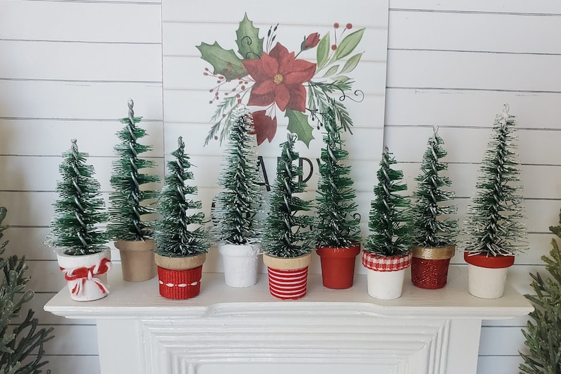 May include: Seven miniature artificial Christmas trees in pots with red and white ribbon accents. The trees are green and have a snowy white finish. The trees are arranged on a white mantel.