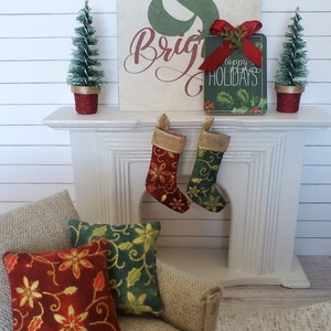 May include: A white fireplace mantel with two red and green stockings hanging from it. There are two small artificial Christmas trees in red pots on either side of the sign that reads "Merry & Bright" with a green sign that reads "Happy Holidays" in front of it. There are two red and green pillows on a beige couch.
