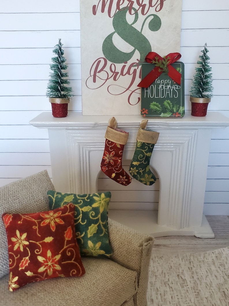May include: A white fireplace mantel with two red and green stockings hanging from it. There are two small artificial Christmas trees in red pots on either side of the sign that reads "Merry & Bright" with a green sign that reads "Happy Holidays" in front of it. There are two red and green pillows on a beige couch.