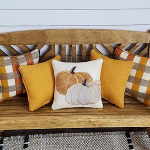 May include: A wooden bench with five pillows. Two pillows are a plaid pattern in orange, brown, and white. Two pillows are solid yellow. One pillow is white with an embroidered design of two pumpkins.