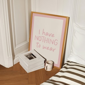 Può includere: Una stampa artistica incorniciata con la scritta "I have NOTHING to wear" in lettere rosa. La stampa è in una cornice dorata con un bordo rosa. Una pila di riviste e una tazza piccola sono sul pavimento accanto alla stampa.