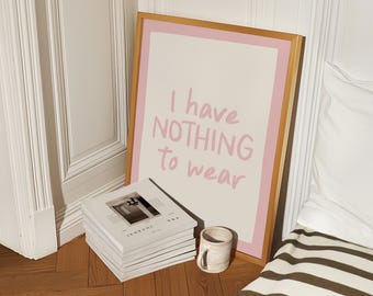 May include: A framed art print with the text "I have NOTHING to wear" in pink lettering. The print is in a gold frame with a pink border. A stack of journals and a small mug sit on the floor next to the print.