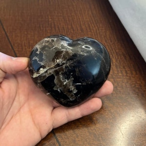May include: A black heart-shaped obsidian crystal with a shimmering, iridescent effect. The crystal is smooth and polished, with a natural, organic shape.