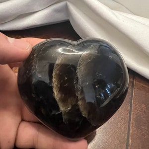 May include: A heart-shaped black obsidian crystal with a natural, cloudy, gray pattern inside. The crystal is polished and smooth.