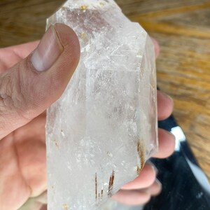 May include: A clear quartz crystal with a point, featuring thin, reddish-brown inclusions. The crystal is approximately 4 inches tall and has a natural, rough texture.