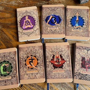 May include: A collection of seven notebooks with mandala designs and yoga poses. Each notebook features a different color scheme and a corresponding chakra symbol. The notebooks are on a wooden surface.
