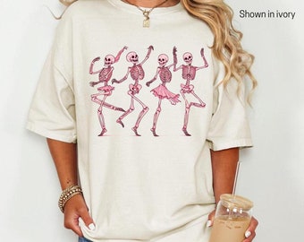 Puede incluir: Una camiseta blanca con un gráfico de cinco esqueletos rosas bailando. El texto "Shown in ivory" está en la esquina superior derecha de la imagen.