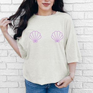 Può includere: T-shirt color crema con due grafiche di conchiglie color lavanda. La maglietta ha una vestibilità ampia e maniche corte. Il testo "chill vibes" è stampato sotto la conchiglia sinistra.