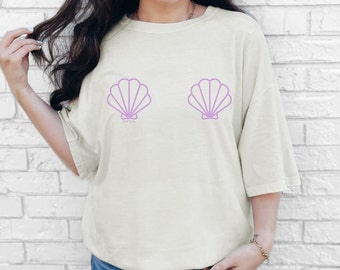 Può includere: T-shirt color crema con due grafiche di conchiglie color lavanda. La maglietta ha una vestibilità ampia e maniche corte. Il testo "chill vibes" è stampato sotto la conchiglia sinistra.