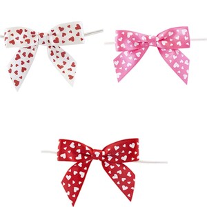 May include: Three gift bows with wire stems. The bows are made of satin ribbon with a white background and red hearts pattern. The bows are in white, pink, and red.