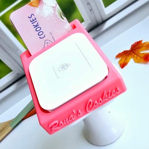 May include: A pink stand with the words "Rona's Cookies" printed on it. A white Square reader is in the stand. A business card with the words "Jore LLC Cookies" is in the stand.