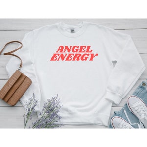 Puede incluir: Sudadera blanca con las palabras "ANGEL ENERGY" impresas en rojo en la parte delantera.