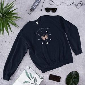 Puede incluir: Una sudadera de color azul marino con un gr&aacute;fico de una mariposa y fases lunares. El texto "ALLOW TRANSFORMATION" est&aacute; impreso en un c&iacute;rculo alrededor de la mariposa.