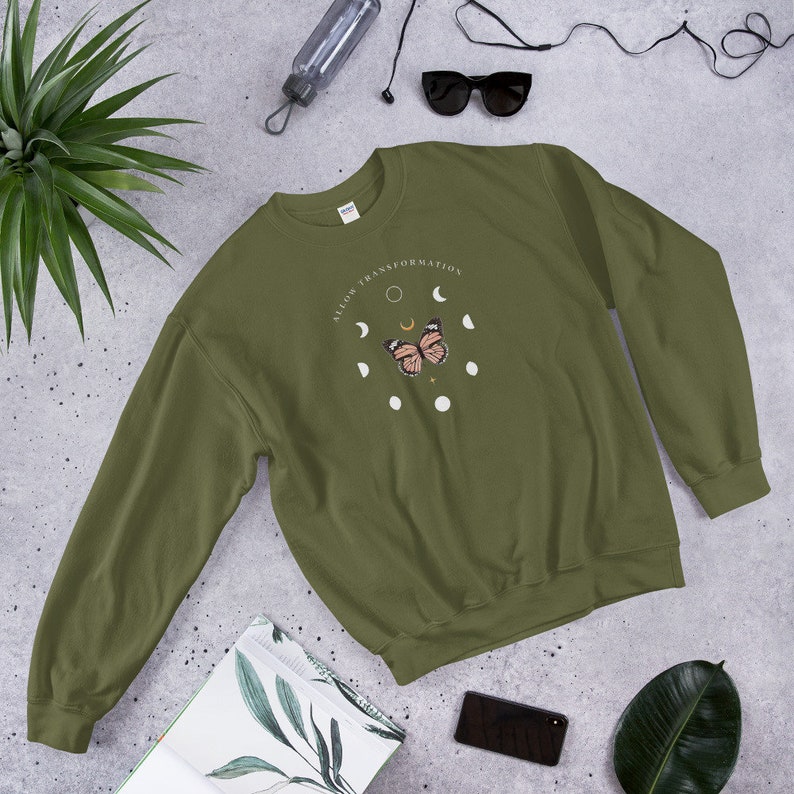 Puede incluir: Una sudadera verde con un gr&aacute;fico de mariposa y fases lunares. El texto "Allow Transformation" est&aacute; impreso en la sudadera.