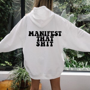 May include: White hooded sweatshirt with the text "MANIFEST THAT SHIT" printed in black on the back.