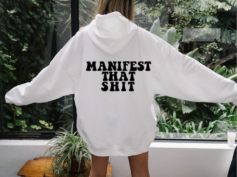May include: White hooded sweatshirt with the text "MANIFEST THAT SHIT" printed in black on the back.
