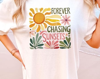 May include: Ivory-colored t-shirt with a retro graphic design. The design features a sun, flowers, and the words "FOREVER CHASING SUNSETS" in a colorful, vintage style. The shirt is a relaxed fit.
