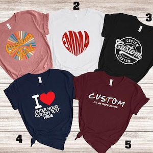 May include: Five t-shirts with custom designs. One is dusty rose with "Custom Text" in a retro design. Another is white with "EMMA" in a heart shape. A black shirt has "Custom" in a circle. A navy shirt says "I ❤️ ENTER YOUR CUSTOM TEXT HERE". A maroon shirt says "CUSTOM I'll be there for you".