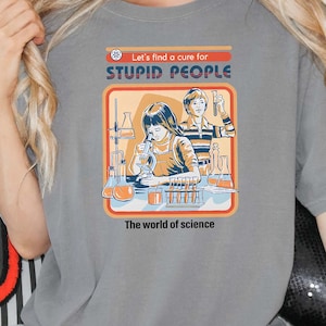 May include: A gray t-shirt with a graphic that reads "Let's find a cure for STUPID PEOPLE." The graphic depicts a vintage science lab scene with beakers and lab equipment. The words "The world of science" are below the graphic.