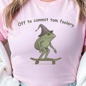 May include: Pink t-shirt with the phrase "Off to commit tom foolery" and a graphic of a green frog wearing a wizard hat and riding a skateboard. The word "Pink" is written in a pink font in the upper right corner.