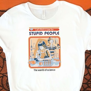 May include: White t-shirt with a retro-style graphic featuring two children in a science lab, with the text "Let's find a cure for STUPID PEOPLE" and "The world of science". The shirt is displayed on an orange background with Halloween decorations.