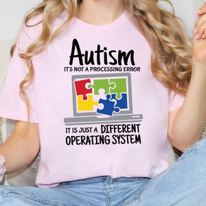 May include: A light pink t-shirt with the word "Autism" in black script above the text "It's not a processing error." Below is a laptop graphic with puzzle pieces. The text "It is just a different operating system" is below the laptop graphic.