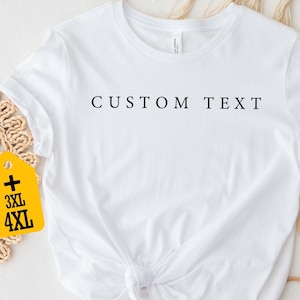 May include: White t-shirt with "CUSTOM TEXT" printed in black. The shirt is knotted at the waist. A yellow tag with "3XL" and "4XL" is attached. The shirt is displayed on a neutral background with decorative elements.