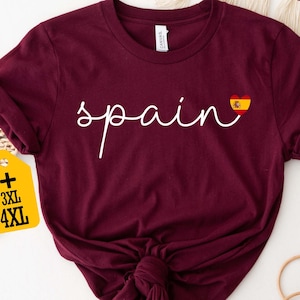 May include: Maroon t-shirt with the word "spain" in white script and a heart with the Spanish flag. The shirt has rolled-up sleeves and a knot at the bottom. A yellow tag indicates sizes 3XL and 4XL.