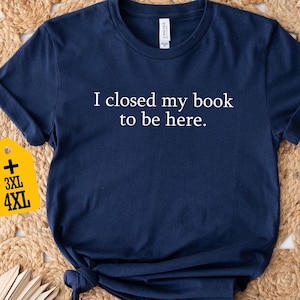 May include: Navy blue t-shirt with the text "I closed my book to be here." A yellow tag indicates 3XL and 4XL sizes. The shirt is displayed on a woven, beige surface.