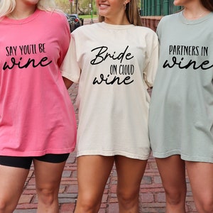 May include: Three women wearing oversized t-shirts with wine-themed phrases. The shirts are in pink, cream, and sage green. The pink shirt says "Say You'll Be Wine", the cream shirt says "Bride on Cloud Wine", and the green shirt says "Partners in Wine".