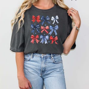 K&ouml;nnte beinhalten: Ein dunkelgraues T-Shirt mit einem Muster aus roten, wei&szlig;en und blauen Schleifen. Die Schleifen sind in einem Raster angeordnet und haben einen Aquarell-Effekt. Das Shirt hat eine lockere Passform und kurze &Auml;rmel.