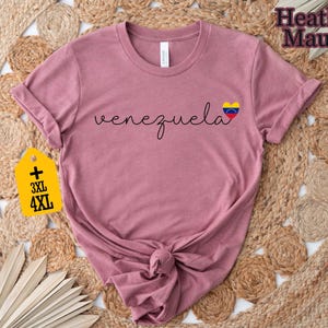 May include: Heather mauve t-shirt with the word "venezuela" in cursive and a heart-shaped flag graphic. The shirt has rolled-up sleeves and a knotted hem. A yellow tag indicates 3XL and 4XL sizes.