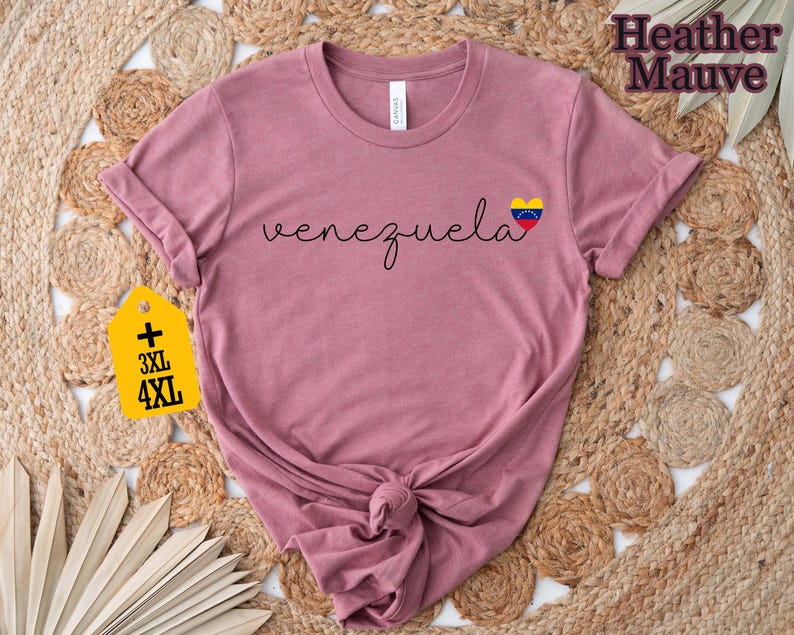 May include: Heather mauve t-shirt with the word "venezuela" in cursive and a heart-shaped flag graphic. The shirt has rolled-up sleeves and a knotted hem. A yellow tag indicates 3XL and 4XL sizes.
