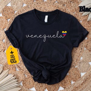 May include: Black t-shirt with the word "Venezuela" in a cursive font and a small heart-shaped Venezuelan flag graphic. The shirt has rolled-up sleeves and a knot at the bottom. A yellow tag indicates sizes 3XL and 4XL.