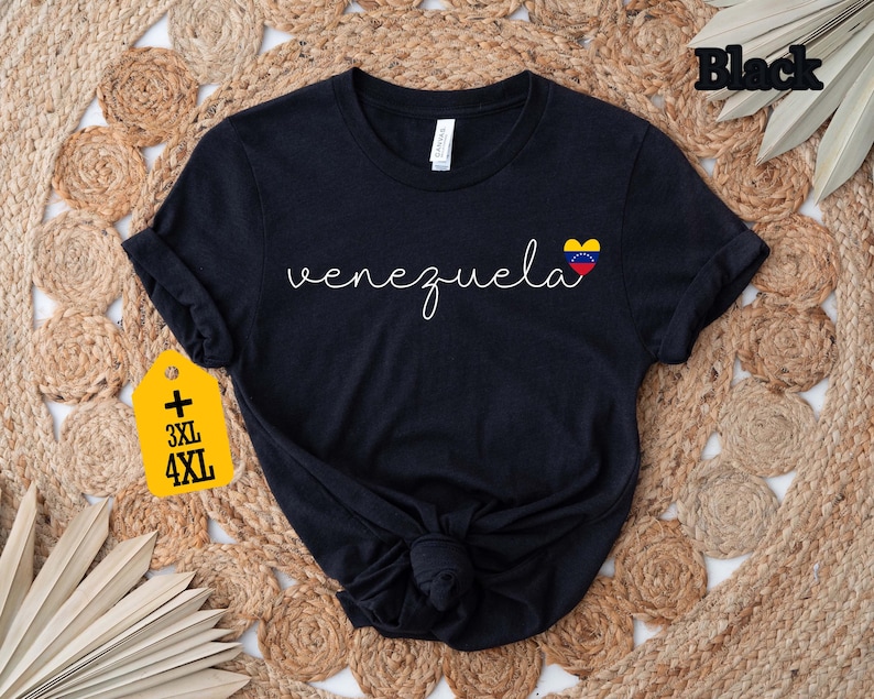 May include: Black t-shirt with the word "Venezuela" in a cursive font and a small heart-shaped Venezuelan flag graphic. The shirt has rolled-up sleeves and a knot at the bottom. A yellow tag indicates sizes 3XL and 4XL.