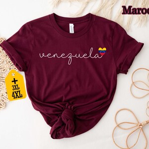 May include: A maroon t-shirt with the word "venezuela" in white script, and a heart-shaped flag graphic. The shirt has rolled-up sleeves and a knot at the bottom. A yellow tag with "3XL" and "4XL" is attached.