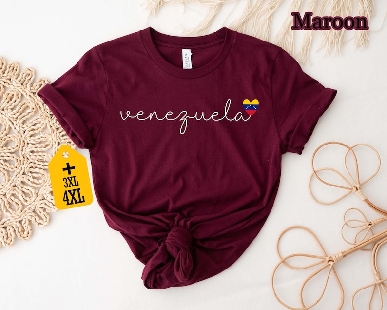 May include: A maroon t-shirt with the word "venezuela" in white script, and a heart-shaped flag graphic. The shirt has rolled-up sleeves and a knot at the bottom. A yellow tag with "3XL" and "4XL" is attached.